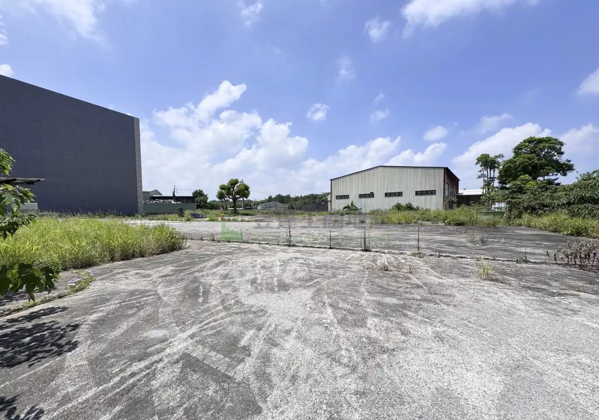 中壢園區外丁建土地一帶現況示意：平整的水泥鋪面空地，遠處是鐵皮廠房與一棟多層廠房，晴天