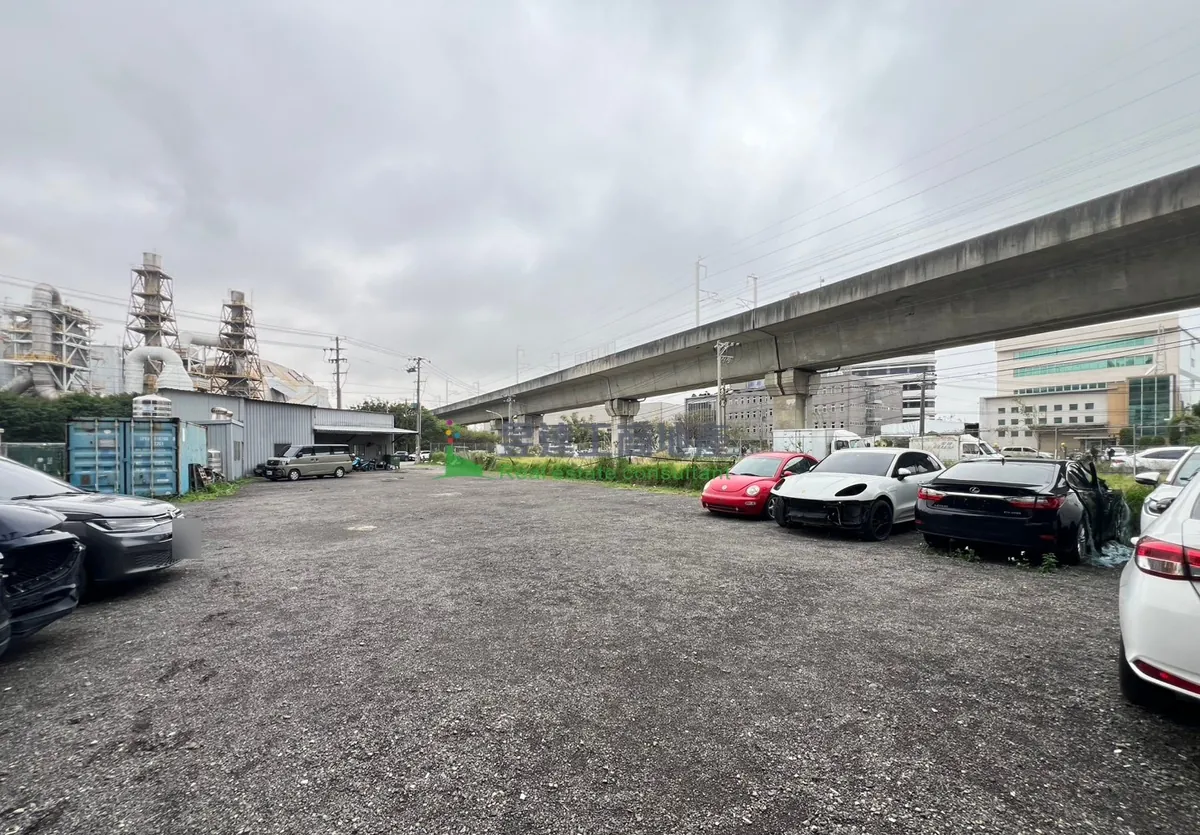 南崁乙種工業區土地一帶現況示意：平整的碎石空地停著幾輛車，左側是鐵皮廠房與貨櫃，遠處有多層廠房，陰天